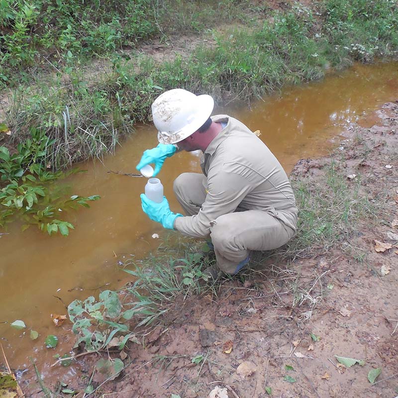 a man wearing a hard hat and gloves taking stormwater samples from a stream.