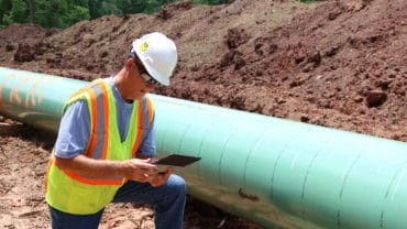a man in a hard hat is kneeling down next to a green pipeline and looking at a tablet.
