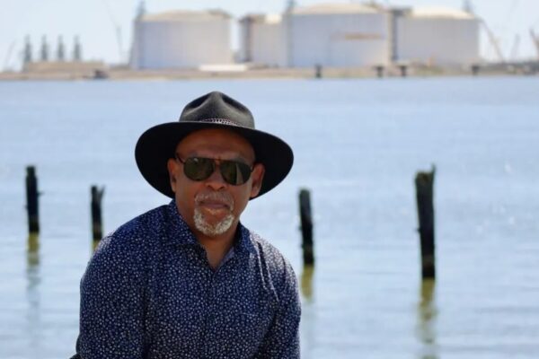 a man wearing a hat and sunglasses is standing in front of a body of water.